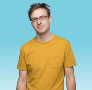 A photo of a white man wearing sunglasses and a friendly smile. He wears a yellow t-shirt and stands in front of a teal background.